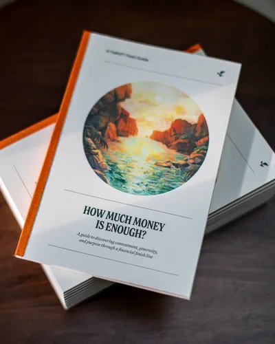 Two stacked copies of the guide “How Much Money Is Enough?”, with a painted seascape on the cover, on a dark wooden table