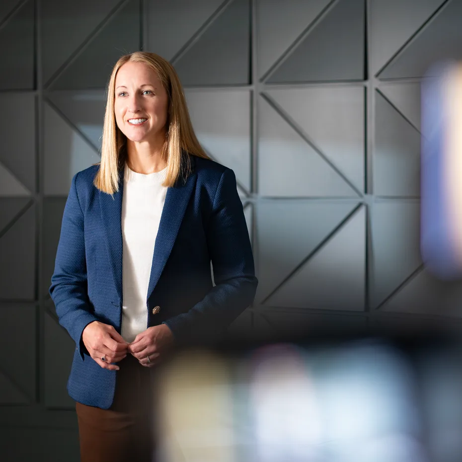 Smiling woman with long blonde hair in a navy blazer standing in front of a dark geometric wall, with blurred studio lights in the foreground