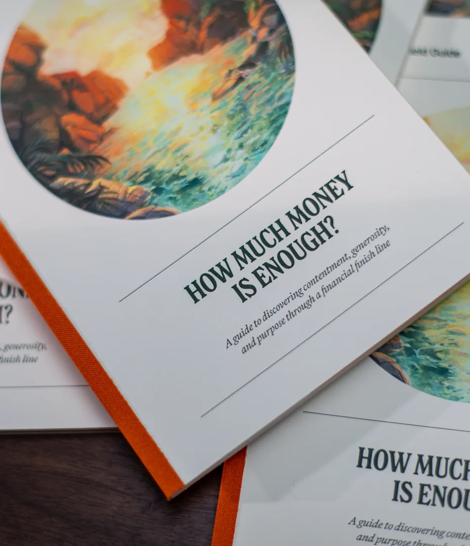 Copies of the guide “How Much Money Is Enough?” with painted seascape covers and orange spines, spread across a table