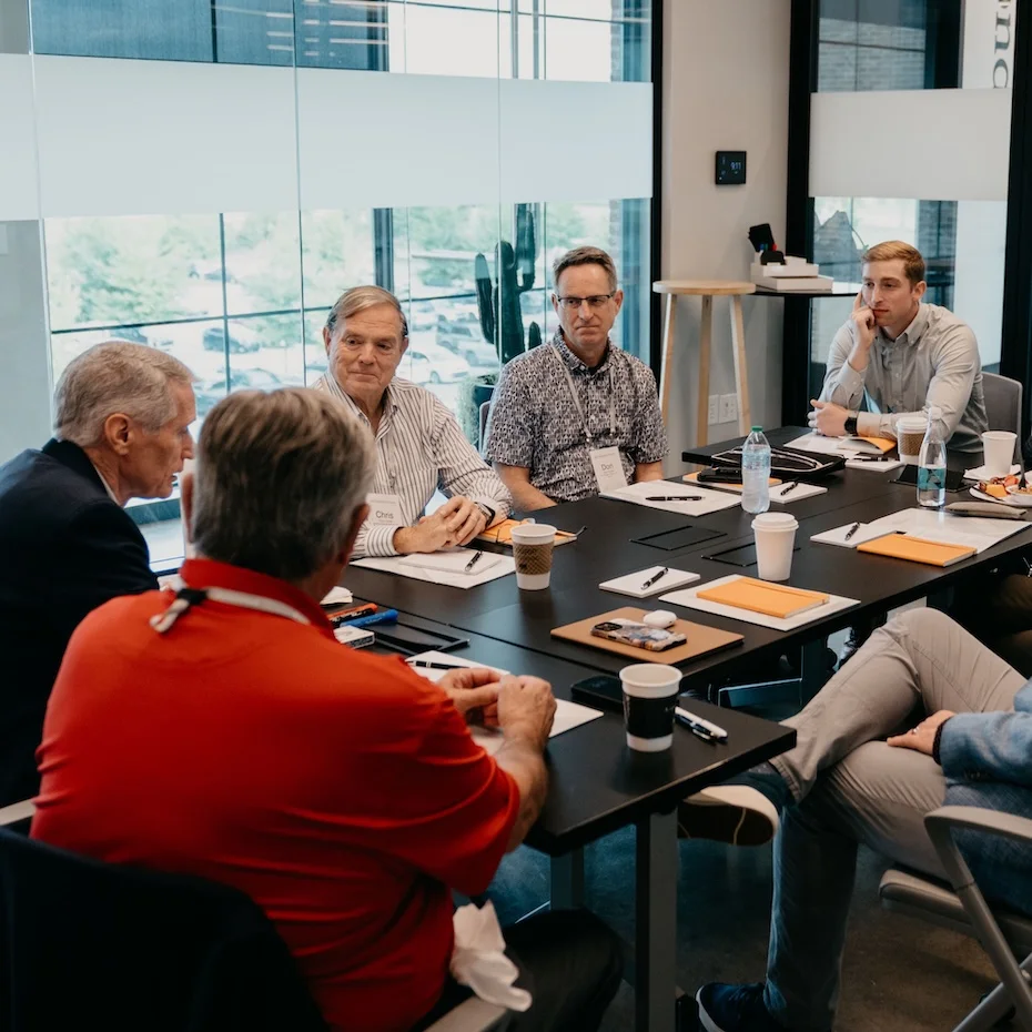 Seven men seated around a conference table engaged in a group discussion during the Kingdom Advisors Champions Forum in a modern, glass-walled meeting room.