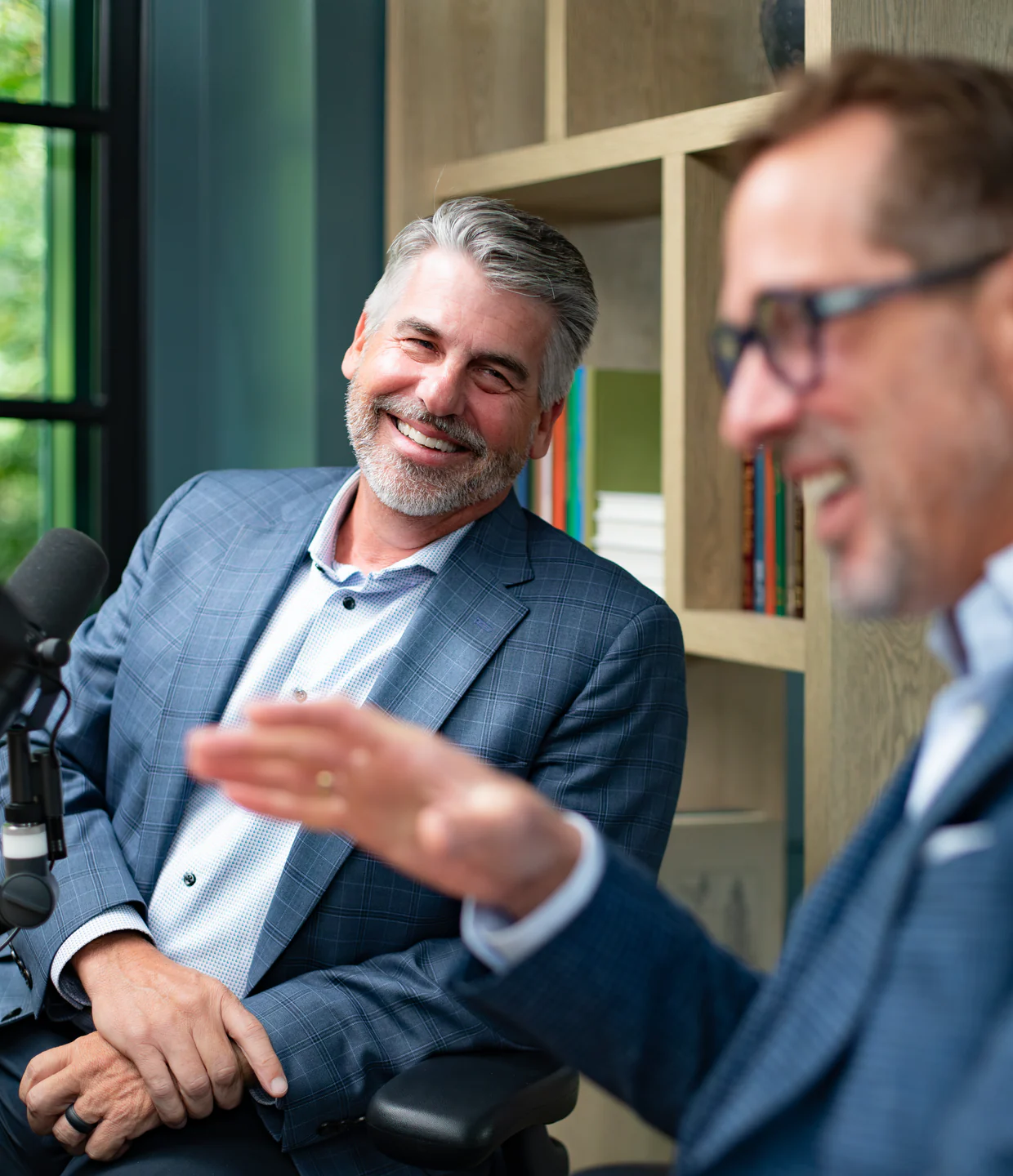 Grey-haired, bearded man in a blue check blazer laughing with a man in glasses beside a podcast microphone, bookshelves behind them