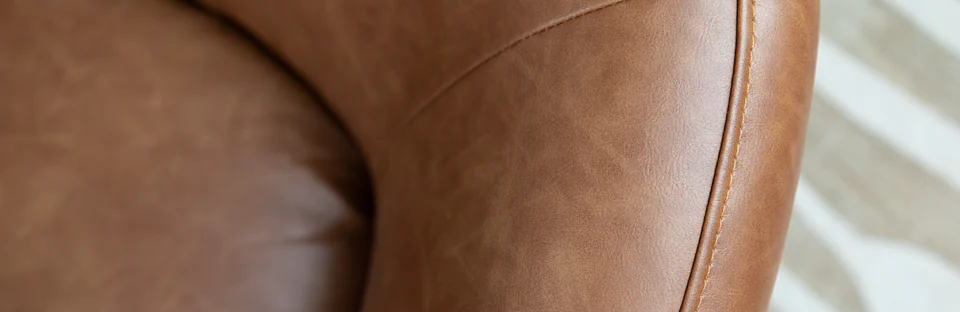 Close-up of the stitched, curved arm of a brown leather armchair beside a cream patterned rug