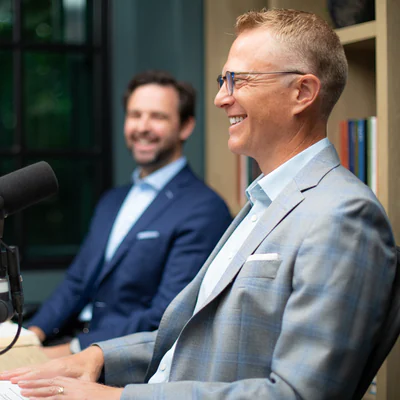 Smiling man with glasses in a grey check suit seated at a studio microphone, with a second smiling man in a navy suit behind him and bookshelves in the background