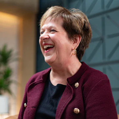 Laughing woman with short auburn hair in a burgundy jacket with gold buttons, seated in front of a teal geometric wall